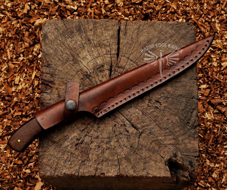 May include: A handcrafted hunting knife with a dark wooden handle and a brown leather sheath. The sheath features decorative stitching and a strap with a button closure. The knife is resting on a wooden block.
