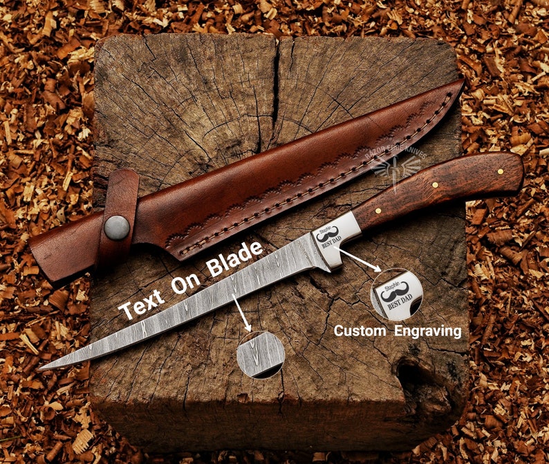 May include: A Damascus steel fillet knife with a wooden handle and a brown leather sheath. The blade has a pointed tip and is engraved with the text "Text On Blade". The handle has brass rivets. The knife is on a wooden surface.