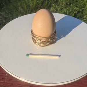 May include: A light brown egg in a gold-colored wire egg cup on a white plate. A green-tipped matchstick rests on the plate. The scene is lit by sunlight, casting a shadow.