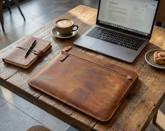 Peut inclure: Une housse d'ordinateur portable en cuir marron avec fermeture éclair, un carnet et un stylo sont posés sur une table en bois. Un ordinateur portable et un café avec du latte art sont également sur la table. Une pâtisserie est sur une petite assiette.
