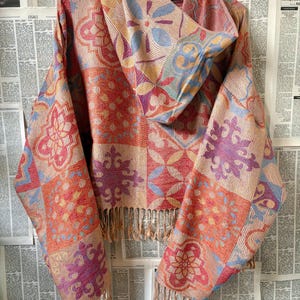 Op de afbeelding: Een kleurrijke sjaal met capuchon en patroon, hangend aan een witte hanger. De sjaal heeft een herhalend bloemmotief in oranje, roze, paars en blauw. De onderkant heeft franjes.