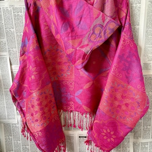 Op de afbeelding: Een felroze sjaal met ingewikkelde bloemen- en geometrische patronen. De sjaal heeft een gefranjerde rand en is over een hanger gedrapeerd. Het kleurenpalet omvat tinten roze, paars en oranje, waardoor het een visueel aantrekkelijk accessoire is.