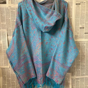 Op de afbeelding: Een turquoise poncho met capuchon en een paisley patroon in roze en paars. De poncho heeft een franje in bijpassende turquoise kleur. Het kledingstuk hangt aan een zwarte hanger tegen een achtergrond van krantenpagina's.