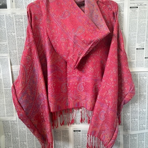 Op de afbeelding: Een felroze sjaal met een paisley patroon en franjes. De sjaal is over een hanger gedrapeerd, wat het ingewikkelde ontwerp en de textuur laat zien. Het kleurenpalet omvat roze en paarse tinten.