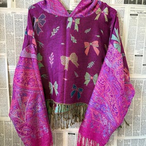 Könnte beinhalten: Ein violetter Kapuzenpullover mit einem sich wiederholenden Muster aus Schleifen und kleinen Zweigen in Rosa-, Blau- und Grüntönen. Das Kleidungsstück hat lange, weite Ärmel mit einem lebhaften Paisley-Muster und einen Saum mit langen Fransen.