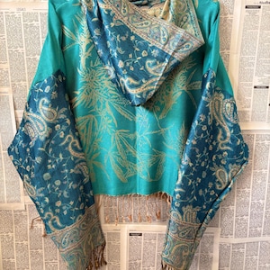 Op de afbeelding: Een turquoise top met lange mouwen, een capuchon en aangehechte sjaalpanelen. Het kledingstuk heeft een herhalend botanisch patroon in een lichtere tint turquoise, terwijl de capuchon en de uiteinden van de sjaal een donkerblauw paisley-motief tonen.