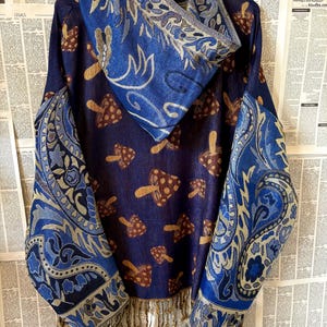 Könnte beinhalten: Eine blaue Kapuzenjacke mit Pilzmuster. Die Ärmel und die Kapuze sind mit Paisley-Mustern in Blau und Beige verziert. Die Jacke hat einen Fransenbesatz und hängt an einem Kleiderbügel.