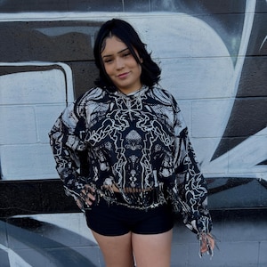 Op de afbeelding: Een zwart-wit patroon cropped hoodie met lange mouwen en een gefranjerde zoom. Het model draagt een zwarte short en kniehoge laarzen. De achtergrond is een zwart-witte graffiti muur.