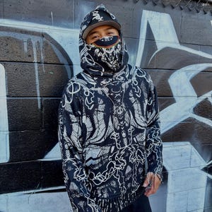 Op de afbeelding: Een persoon draagt een zwart-witte hoodie met patroon, een bijpassend gezichtsmasker en een baseballpet. De hoodie heeft een uniek ontwerp met witte lijnkunst op een zwarte achtergrond. De persoon staat voor een met graffiti bedekte muur.