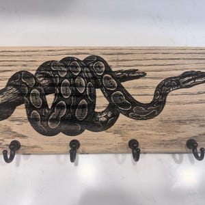 May include: A rectangular light-colored wooden wall-mounted key holder featuring a dark, detailed laser-engraved illustration of a coiled snake resting on a branch. The piece includes four evenly spaced black metal hooks along the bottom edge.