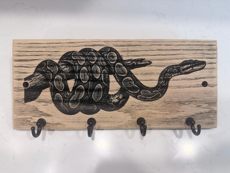 Op de afbeelding: Een rechthoekige, lichtgekleurde houten wand-sleutelhouder met een donkere, gedetailleerde lasergegraveerde illustratie van een opgerolde slang op een tak. Het stuk heeft vier gelijkmatig verdeelde zwarte metalen haken aan de onderkant.