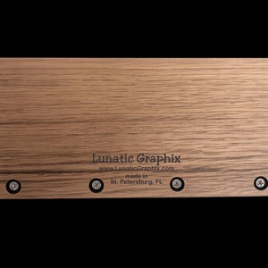 Op de afbeelding: Een rechthoekige lichtbruine houten plaat met een zichtbare houtnerf. De tekst Lunatic Graphix, www.LunaticGraphix.com en made in St. Petersburg, FL is in het zwart gedrukt. Vier zilverkleurige schroeven staan langs de onderrand.