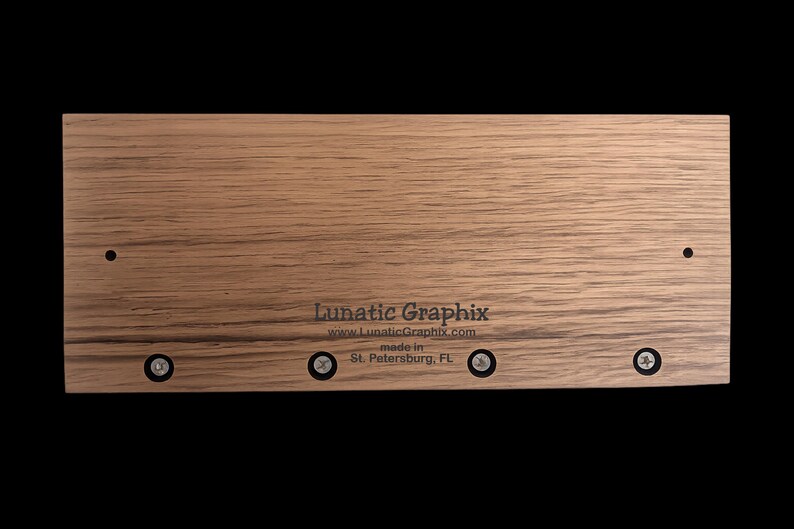 Op de afbeelding: Een rechthoekige lichtbruine houten plaat met een zichtbare houtnerf. De tekst Lunatic Graphix, www.LunaticGraphix.com en made in St. Petersburg, FL is in het zwart gedrukt. Vier zilverkleurige schroeven staan langs de onderrand.