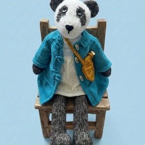 Puede incluir: Muñeca de oso panda tejida a mano, con abrigo azul turquesa, vestido blanco y calcetines grises, sentada en una pequeña silla de madera. El panda tiene marcas en blanco y negro y una pequeña bandolera amarilla. El fondo es azul claro.
