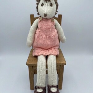 May include: A knitted hedgehog doll, seated on a small wooden chair. The doll has a white face, brown hair, and is wearing a pink dress with wooden button details. It is wearing brown shoes.