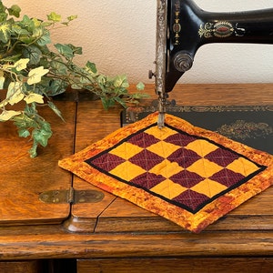 Puede incluir: Primer plano de una máquina de coser vintage con una alfombra de tela a cuadros sobre la mesa. La máquina es negra con detalles dorados y tiene una aguja enhebrada con hilo. La alfombra de tela es un mosaico de cuadrados amarillos y burdeos.