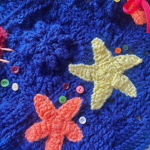 May include: Close-up of a blue crocheted blanket with orange and yellow starfish appliqu&eacute;s. The blanket is decorated with colorful buttons and other crocheted sea creatures. The texture of the yarn is visible.