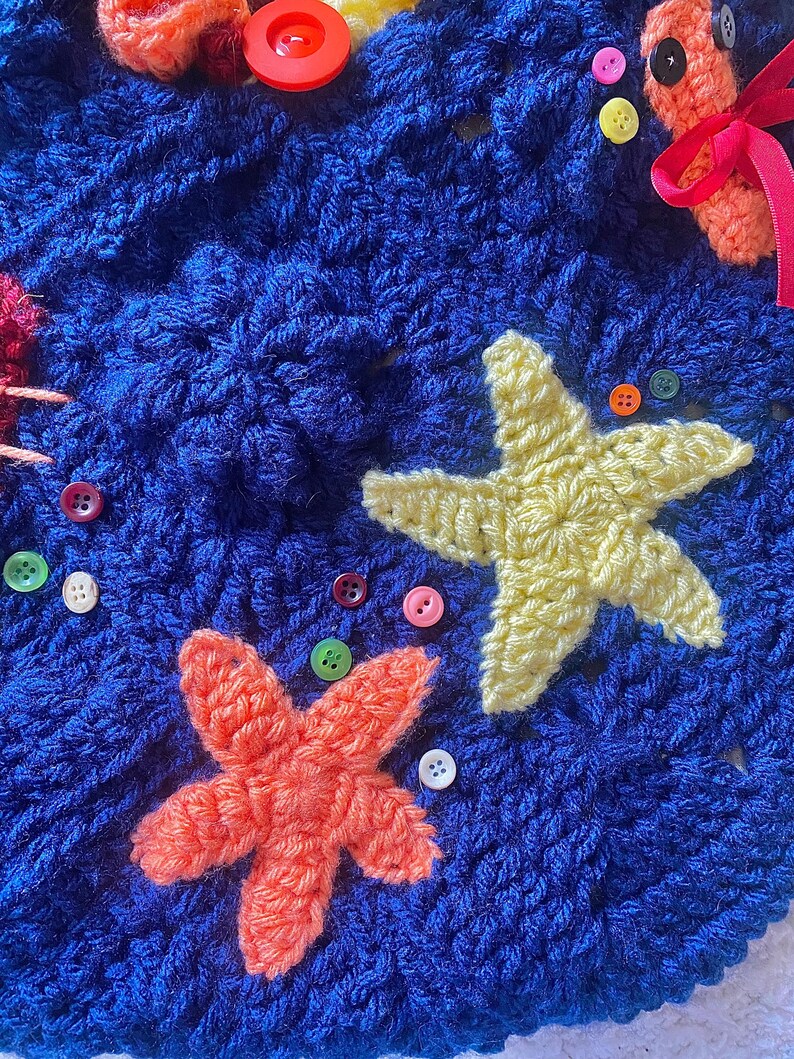 May include: Close-up of a blue crocheted blanket with orange and yellow starfish appliqu&eacute;s. The blanket is decorated with colorful buttons and other crocheted sea creatures. The texture of the yarn is visible.