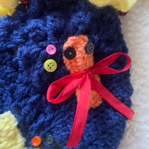 May include: Handmade crochet item in navy blue with yellow, red, and orange accents. Features an orange crochet design with black button eyes and a red ribbon bow. Includes colorful buttons.