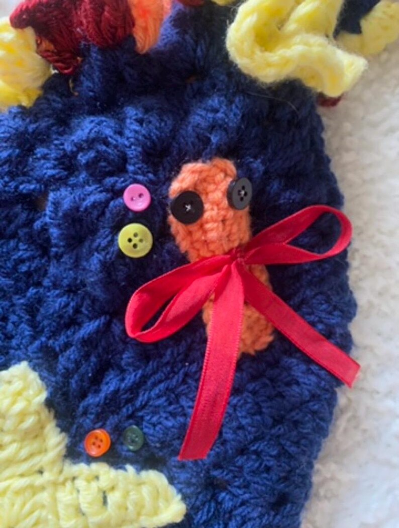 May include: Handmade crochet item in navy blue with yellow, red, and orange accents. Features an orange crochet design with black button eyes and a red ribbon bow. Includes colorful buttons.