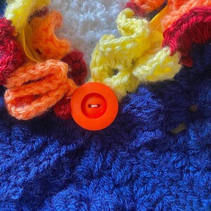 May include: Close-up of a handmade crochet item featuring a vibrant color palette. The design incorporates blue, white, yellow, orange, and red yarn. An orange button is centered on the blue crochet fabric, adding a focal point to the piece.