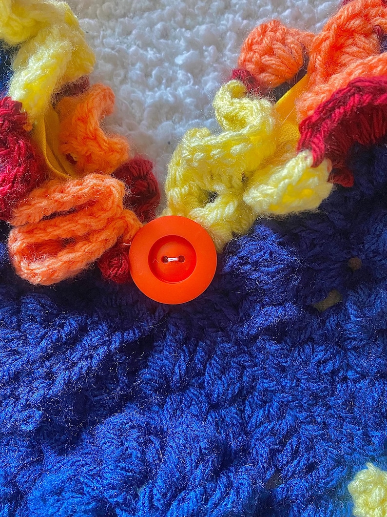 May include: Close-up of a handmade crochet item featuring a vibrant color palette. The design incorporates blue, white, yellow, orange, and red yarn. An orange button is centered on the blue crochet fabric, adding a focal point to the piece.
