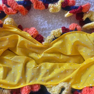 May include: A handmade, colorful crochet piece with a yellow fabric overlay. The crochet features orange, red, yellow, and blue yarn, with a scalloped edge. The yellow fabric has a white polka dot pattern. Buttons are visible at the bottom.