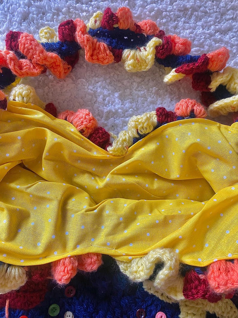 May include: A handmade, colorful crochet piece with a yellow fabric overlay. The crochet features orange, red, yellow, and blue yarn, with a scalloped edge. The yellow fabric has a white polka dot pattern. Buttons are visible at the bottom.