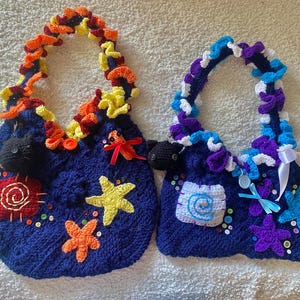 May include: Two handmade, dark blue crochet bags with decorative handles. One has orange, yellow, and red accents, while the other features purple, blue, and white. Both bags are adorned with starfish, buttons, and other embellishments.