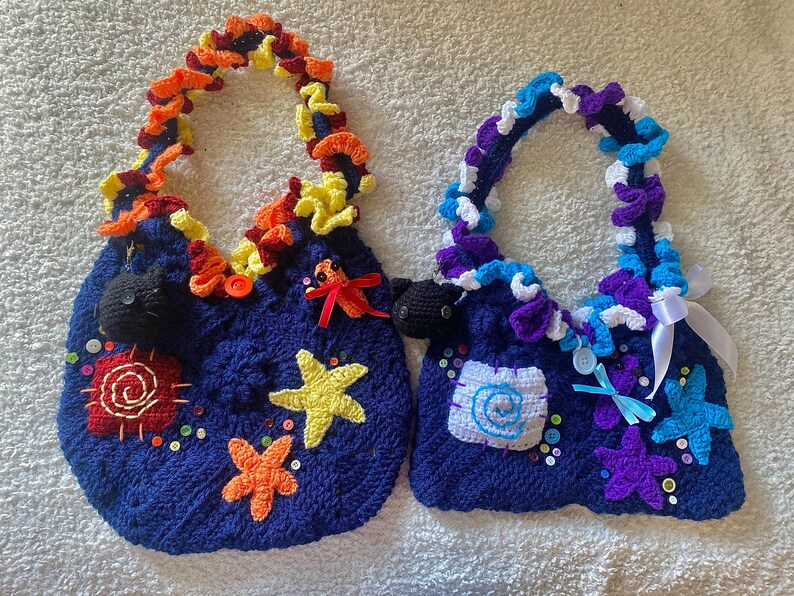 May include: Two handmade, dark blue crochet bags with decorative handles. One has orange, yellow, and red accents, while the other features purple, blue, and white. Both bags are adorned with starfish, buttons, and other embellishments.