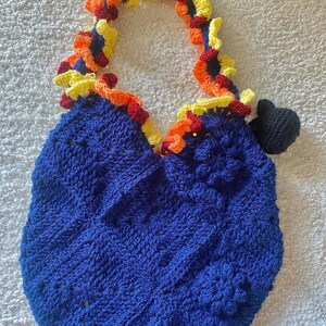 May include: A handmade blue crochet bag with a decorative handle and trim. The handle and trim feature a colorful design with yellow, orange, and red accents. A small black crochet detail is attached to the side of the bag.