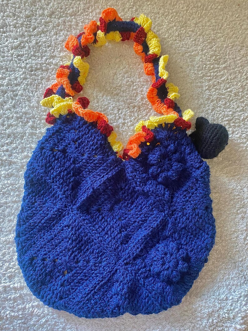 May include: A handmade blue crochet bag with a decorative handle and trim. The handle and trim feature a colorful design with yellow, orange, and red accents. A small black crochet detail is attached to the side of the bag.