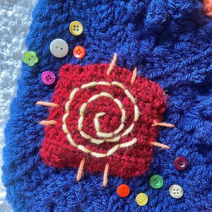 May include: A close-up of a blue crocheted item with a red, square-shaped applique featuring a cream-colored spiral design. Various colorful buttons, including white, yellow, and pink, are scattered around the applique.