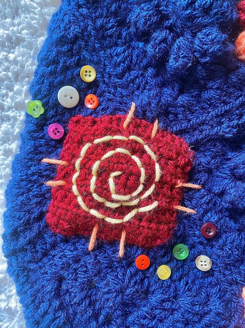 May include: A close-up of a blue crocheted item with a red, square-shaped applique featuring a cream-colored spiral design. Various colorful buttons, including white, yellow, and pink, are scattered around the applique.