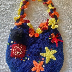 May include: A handmade blue crochet bag with a colorful handle and decorative elements. The bag features a black cat, starfish, and other embellishments. The handle is adorned with yellow, orange, and red crochet details.