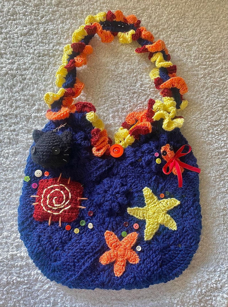 May include: A handmade blue crochet bag with a colorful handle and decorative elements. The bag features a black cat, starfish, and other embellishments. The handle is adorned with yellow, orange, and red crochet details.