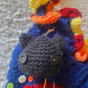 May include: A handmade, crocheted cat-shaped bag in blue, red, yellow, and orange. The bag features a black cat face with button eyes and a variety of colorful buttons. A small, silver-colored charm is attached to the bag.