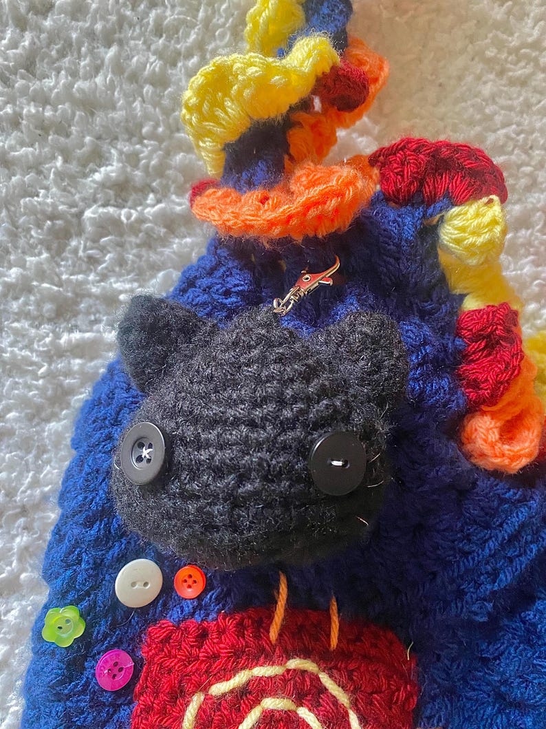 May include: A handmade, crocheted cat-shaped bag in blue, red, yellow, and orange. The bag features a black cat face with button eyes and a variety of colorful buttons. A small, silver-colored charm is attached to the bag.