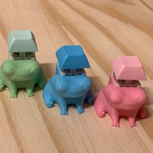 Könnte beinhalten: Drei Froschfiguren, jede in einer anderen Farbe bemalt: grün, blau und rosa. Jeder Frosch hat einen kleinen quadratischen Tastenkappen auf dem Kopf, ebenfalls in einer passenden Farbe.