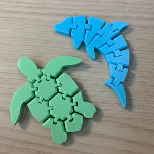 Könnte beinhalten: Eine grüne Plastik-Schildkröte und ein blauer Plastik-Delfin, beide aus ineinandergreifenden Teilen. Die Schildkröte ist nach links und der Delfin nach rechts gerichtet.