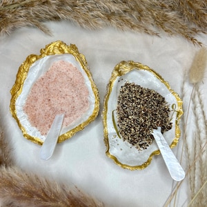 Op de afbeelding: Twee witte keramische oesterschelpen met gouden rand, elk gevuld met een ander kruid. De ene schaal bevat roze Himalaya-zout, de andere zwarte peperkorrels. Elke schaal heeft een witte keramische lepel.