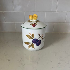 May include: White ceramic jar with a lid, featuring a fruit design. The jar has a green rim and a lid with a yellow fruit-shaped handle. The design includes purple plums and brown leaves. The jar is on a light-colored countertop.