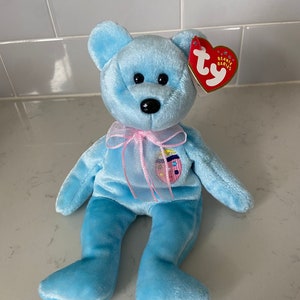 May include: A light blue beanie baby bear with a pink ribbon around its neck. The bear has a small heart-shaped tag that says "Beanie Babies" on it.