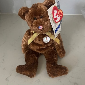 May include: Brown Beanie Baby bear wearing a gold ribbon and a 2002 FIFA World Cup patch. The bear has a small American flag sewn on its nose. The bear is holding a Ty Beanie Baby tag that says "Champion of the Year".