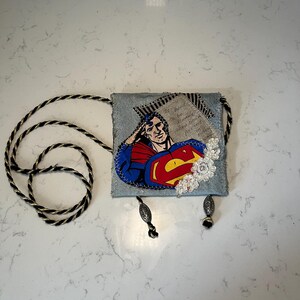 May include: A small denim bag with a Superman graphic, white lace trim, and a black and gold rope strap. The bag has two tassels with metal beads.