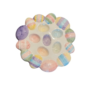 May include: A ceramic Easter egg serving platter. The white platter has a central section with egg-shaped indentations and is surrounded by decorative, pastel-colored eggs. The eggs are painted with various patterns and colors, including pink, blue, yellow, and purple.