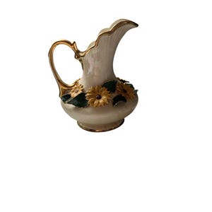 May include: A cream-colored ceramic pitcher with an iridescent finish, featuring a gold-painted handle and rim. The base of the pitcher is decorated with three-dimensional yellow sunflowers with dark brown centers and dark green leaves.