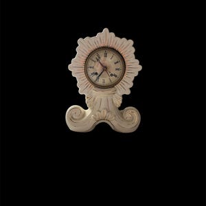May include: A small, off-white decorative clock with a sunburst design around the clock face. The clock has Roman numerals and ornate detailing. The base has a scrolled design. The clock is approximately 6 inches tall.
