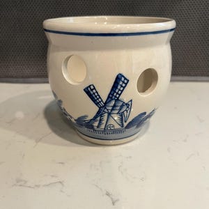 May include: Cream-colored ceramic garlic keeper with a blue windmill design. The keeper has a round shape with three holes and a blue rim. A decorative kitchenware item.