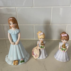 May include: Three porcelain figurines of girls in dresses. The girl on the left is holding a bouquet of flowers and has a number 10 on her dress. The two girls on the right are holding bouquets of flowers and have a number 4 on their dresses.